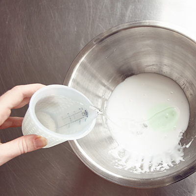 School glue being poured from a measuring cup into a stainless steel mixing bowl to make slime.