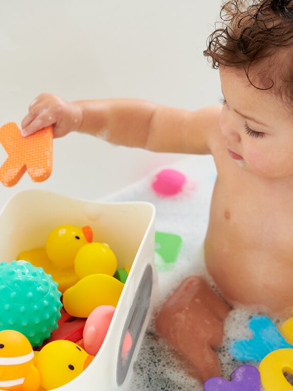 A happy baby at bathtime playing with toys held in a bath toy bin.